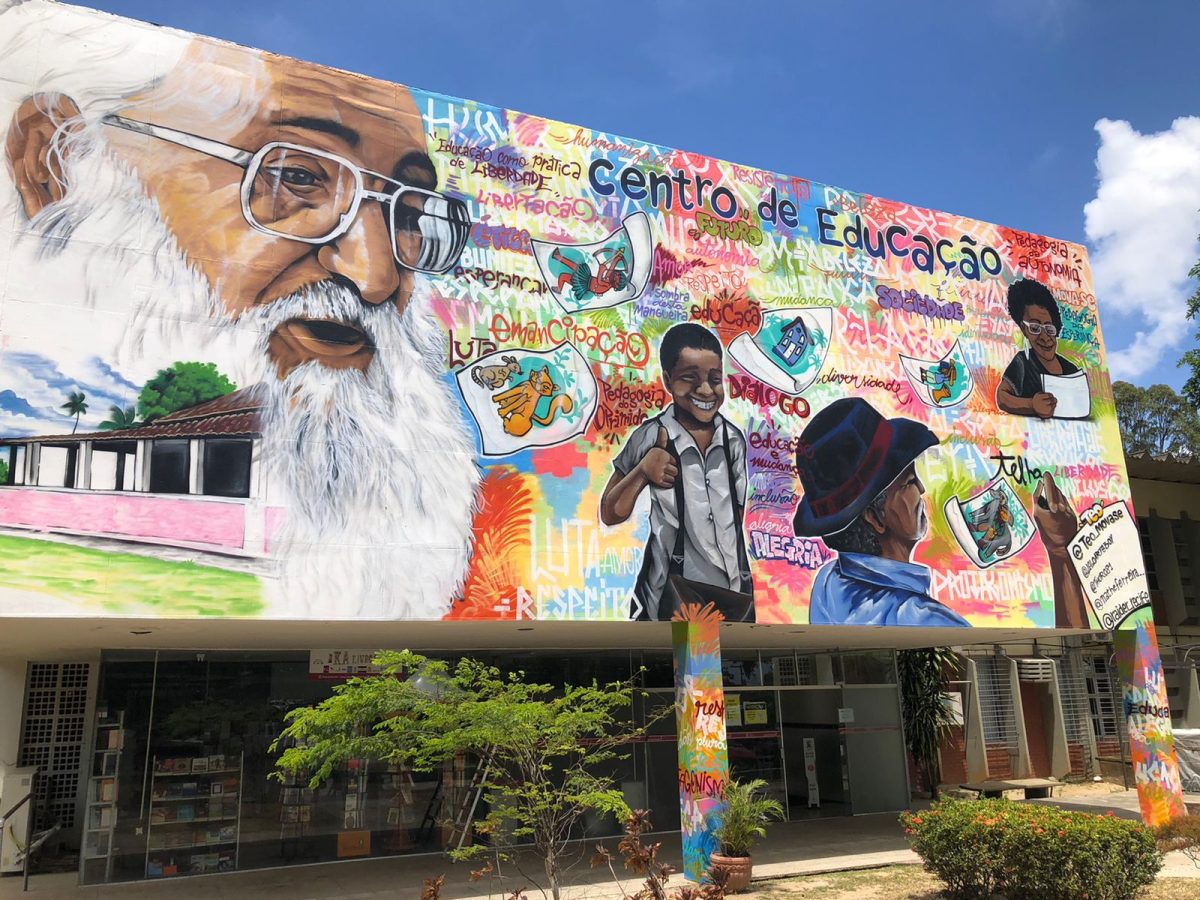 Fotografia do Centro de Educação (CE -UFPE): Sob um toldo com o nome Centro de Educação, Em grafite, está o rosto de Paulo Freire p Ele está  de barba e sorri. À direita, há um garoto e um homem de chapéu que escreve “telha” no mural do CE. Mais acima e à direita do homem, uma senhora negra olha para alguns papéis.